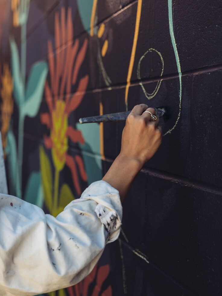Close-up of an artist's hand wearing a ring, using a fine brush to paint white outlines on a dark brick mural featuring floral patterns.